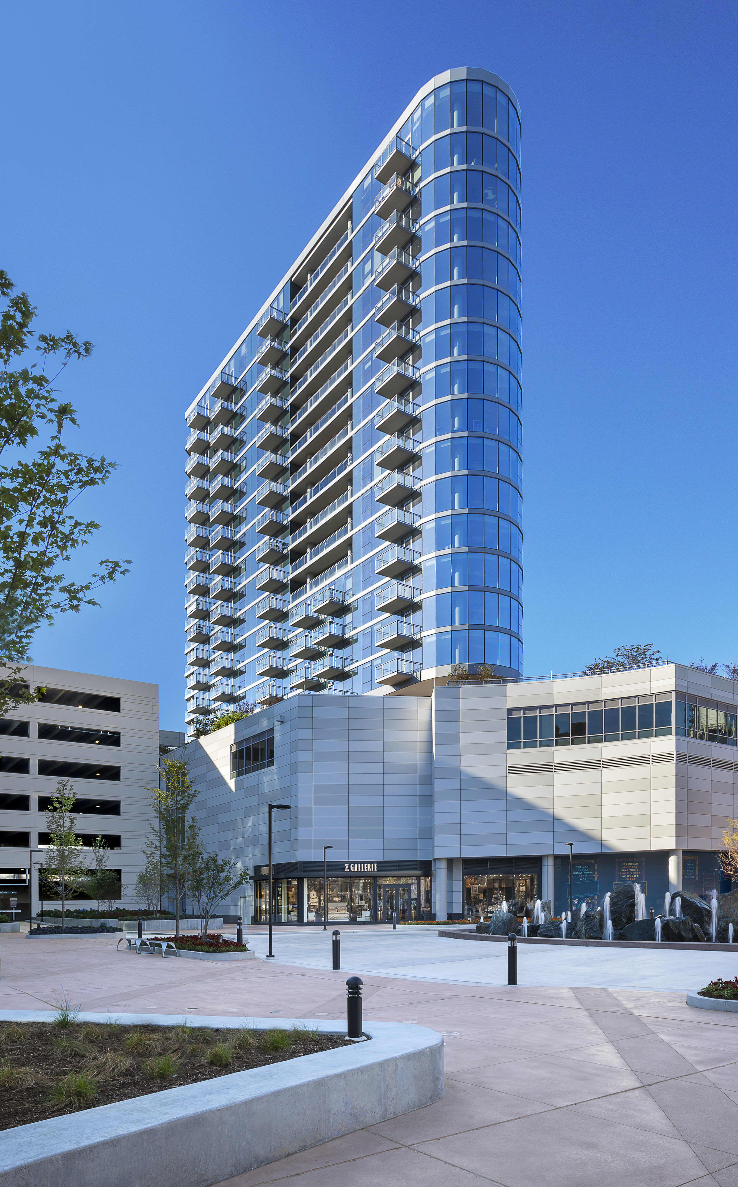 Curved residential tower with blue-tinted glass facade and balconies, white base with retail storefronts, and plaza with landscaping.