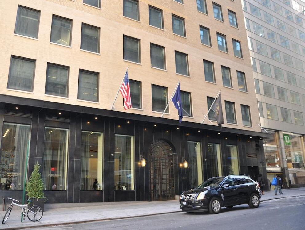 Modern building facade with black storefront, multiple windows, American and state flags, parked SUV, and street-level entrance.