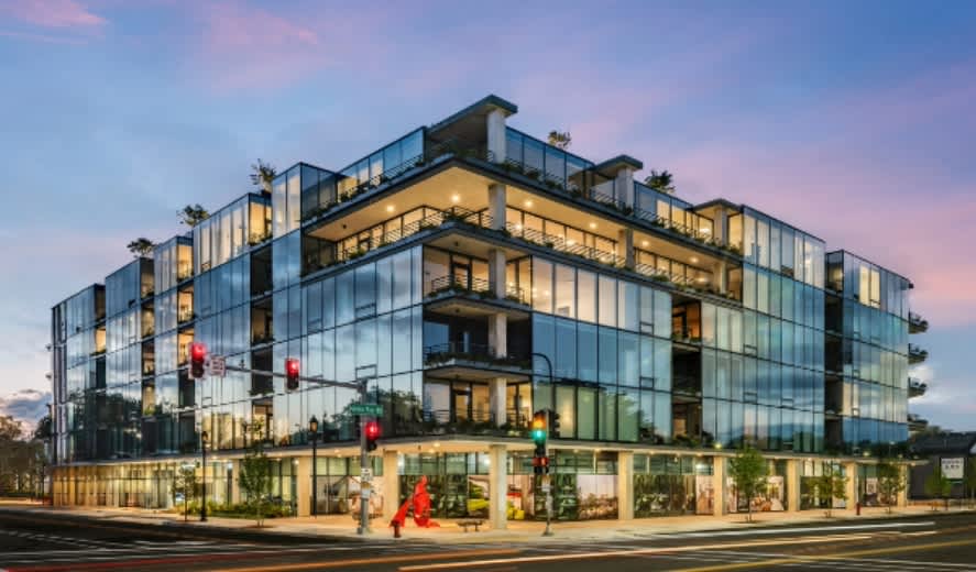 Contemporary four-story apartment building with extensive glass facade, lit interior units, rooftop terraces, and street-level storefronts at dusk.