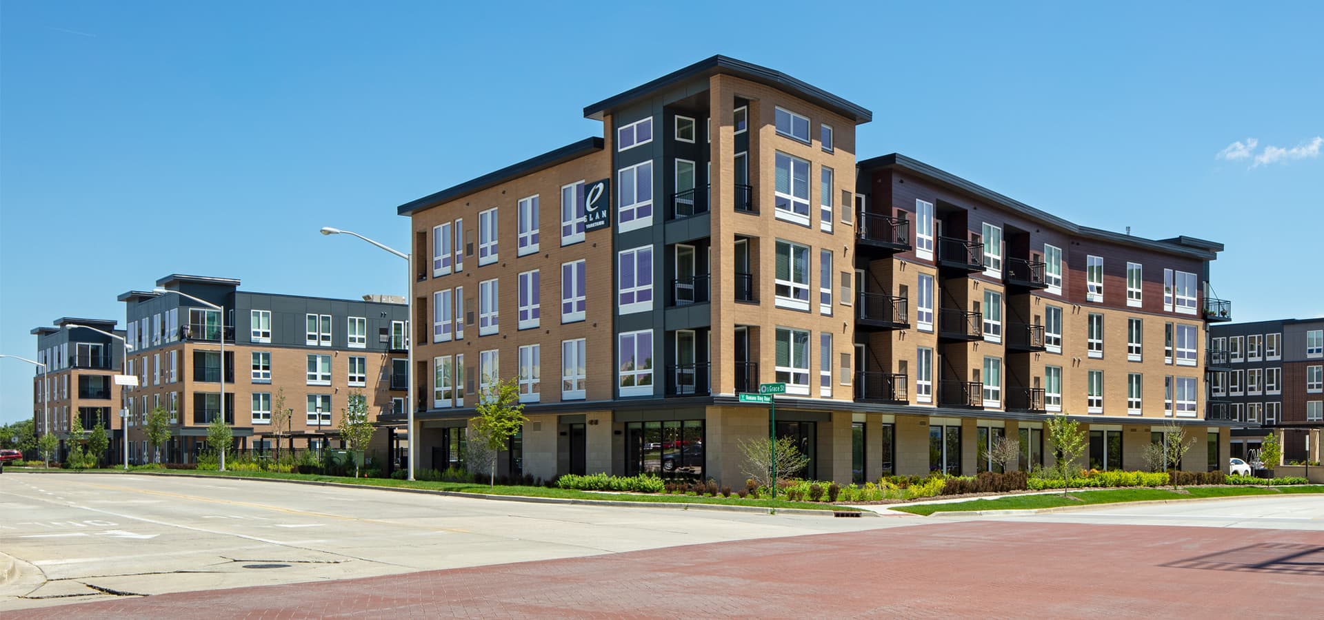 Modern apartment complex with tan and dark brick facades, large windows, ground-floor retail spaces, and landscaped grounds in Lombard.
