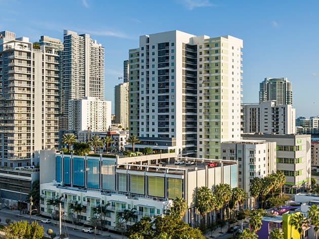 Modern multi-story residential building with green and white facade surrounded by downtown Miami high-rises and palm trees.