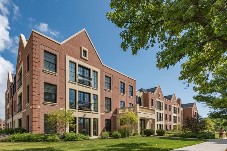Row of red brick townhomes with peaked roofs, balconies, and large windows overlooking landscaped lawn.