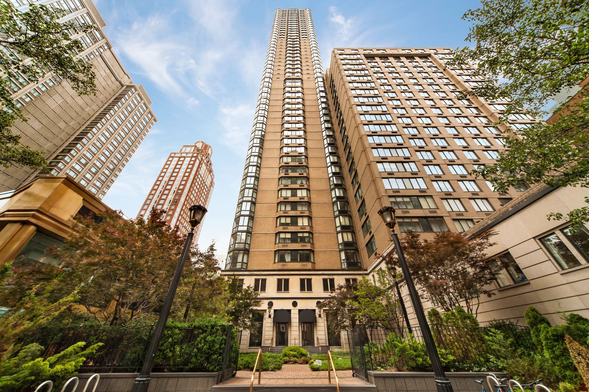 Tall residential tower with brick facade and rows of windows, viewed from landscaped courtyard with mature trees and black lampposts.