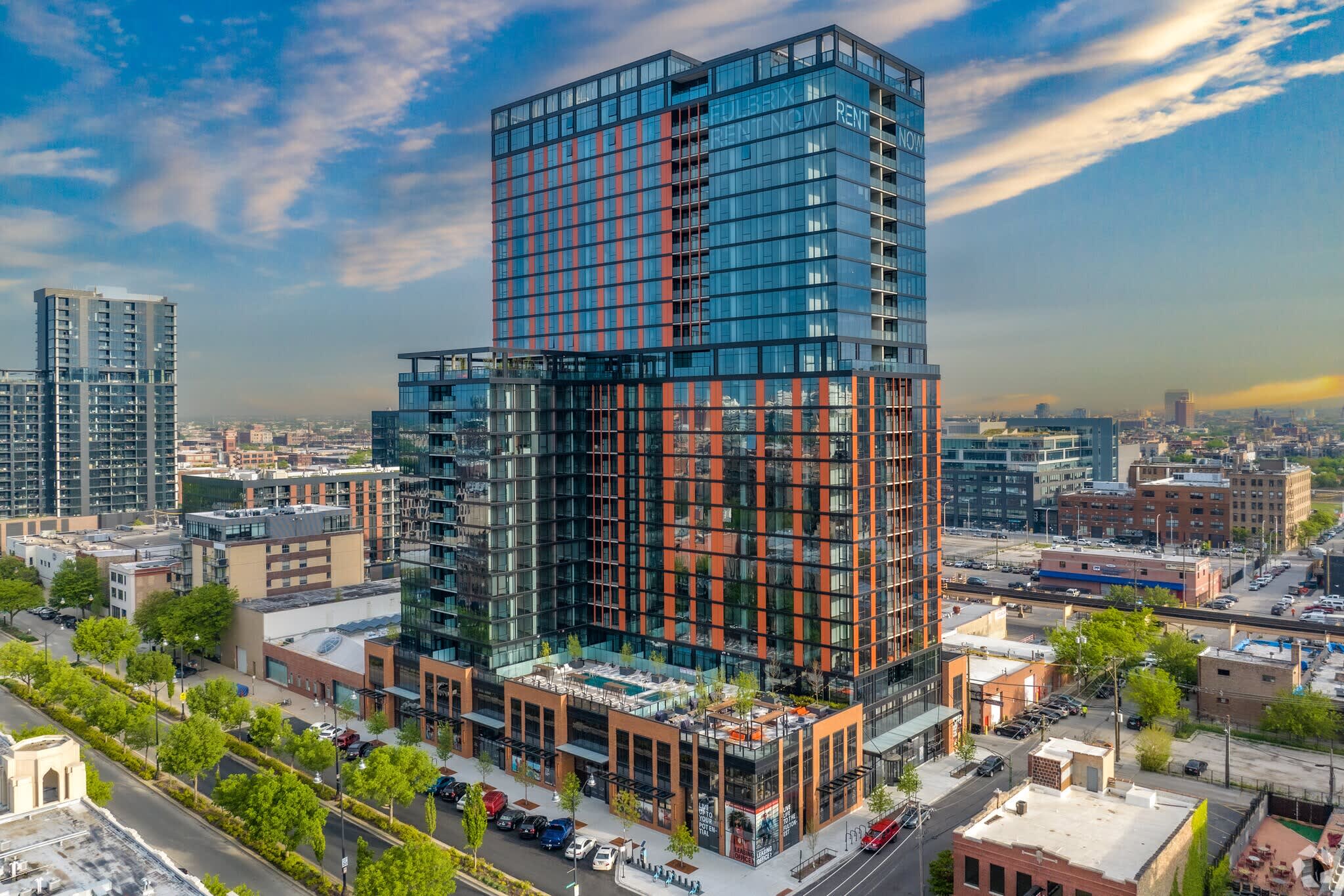 Modern residential tower with red and blue metal facade, glass windows, ground-level retail spaces, and surrounding urban landscape.