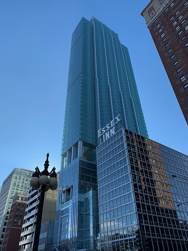 Tall glass residential tower with vertical ribbing and illuminated signage, flanked by older brick buildings and a street lamp against clear blue sky.