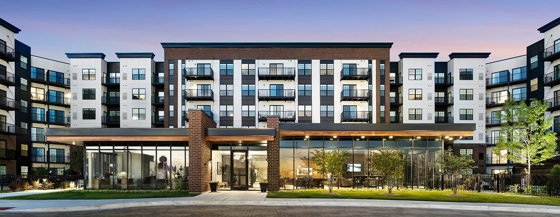 Modern apartment complex with brick and glass ground-level pavilion, multi-story residential towers with balconies, landscaped courtyard with young trees and lawn.