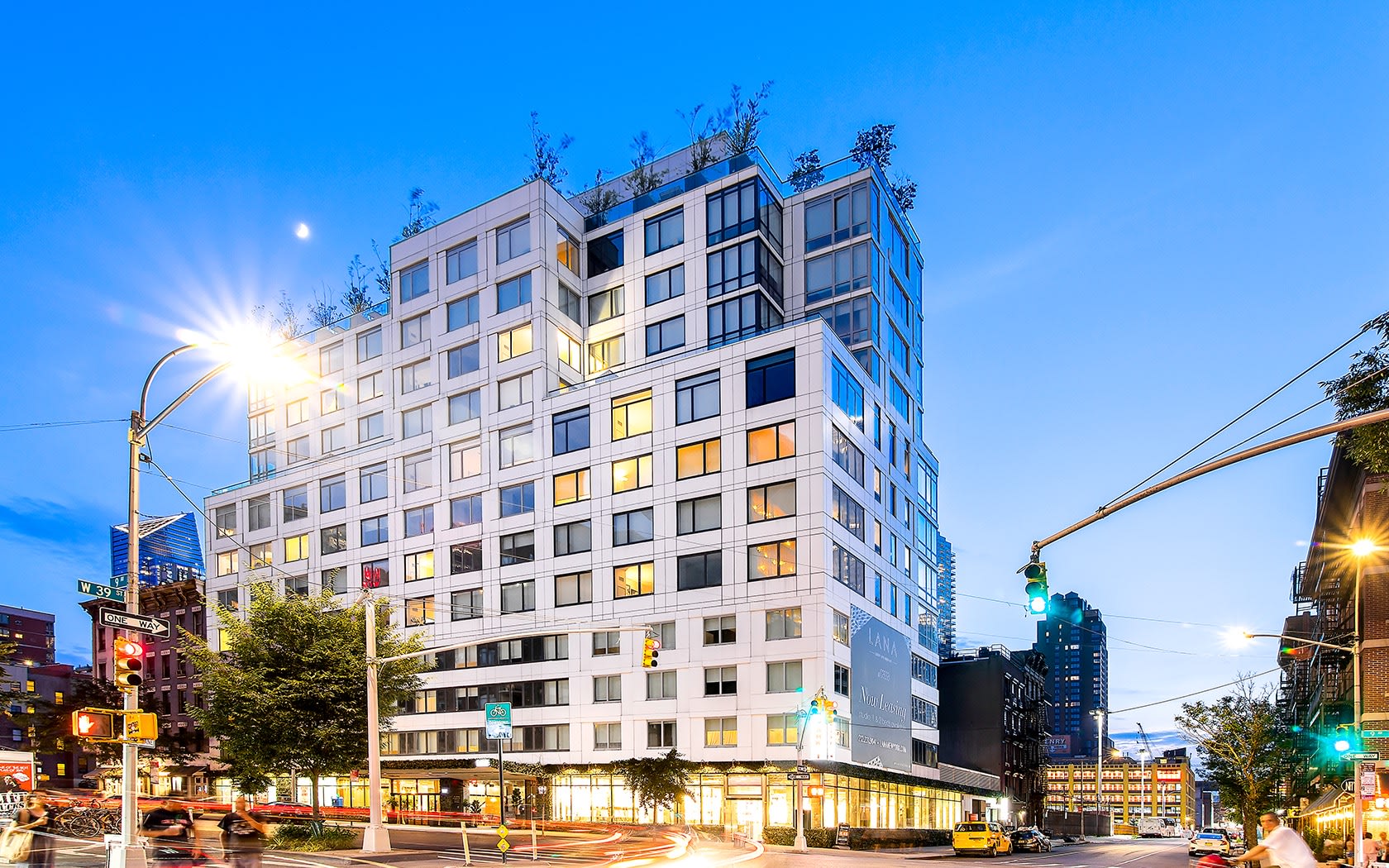Modern multi-story residential building with illuminated windows at dusk, street-level storefronts, and city surroundings.