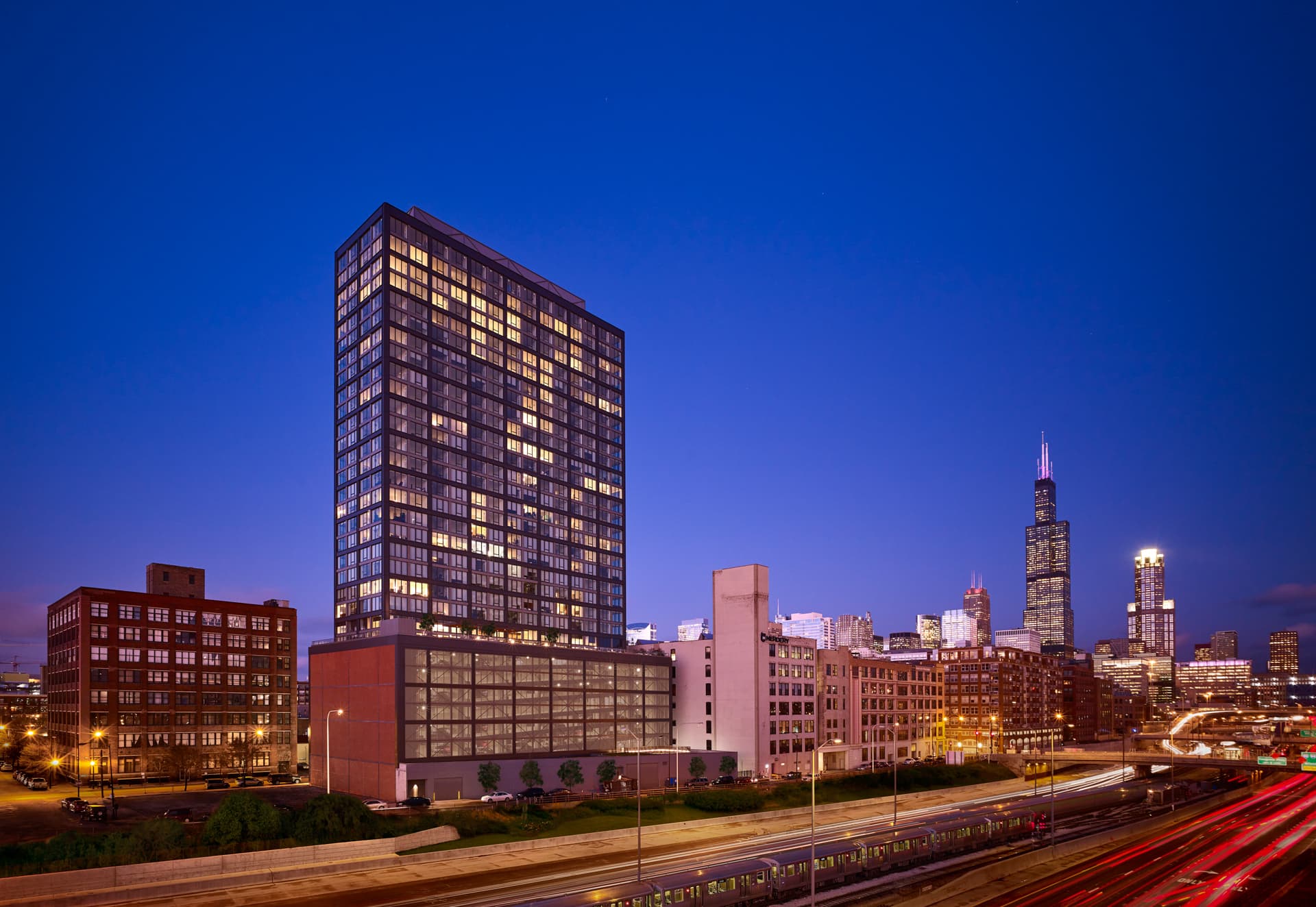 Illuminated high-rise residential tower at dusk with Chicago skyline and elevated train tracks in foreground.