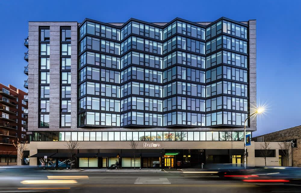 Contemporary multi-story residential building with curved glass balconies, stone and brick facade, ground-floor retail, and clear blue sky.