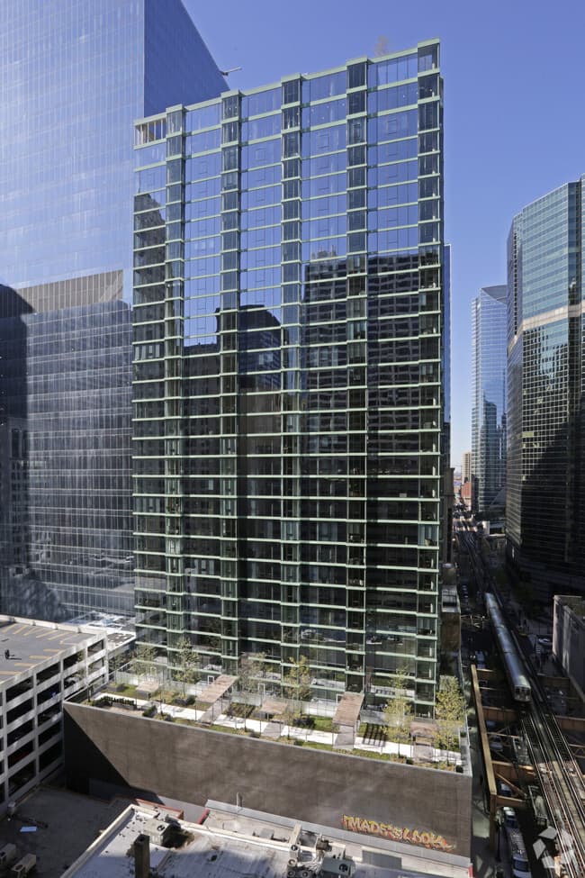 Contemporary high-rise residential building with green-framed glass facade, multiple balconies, and ground-level plaza surrounded by downtown Chicago skyscrapers.