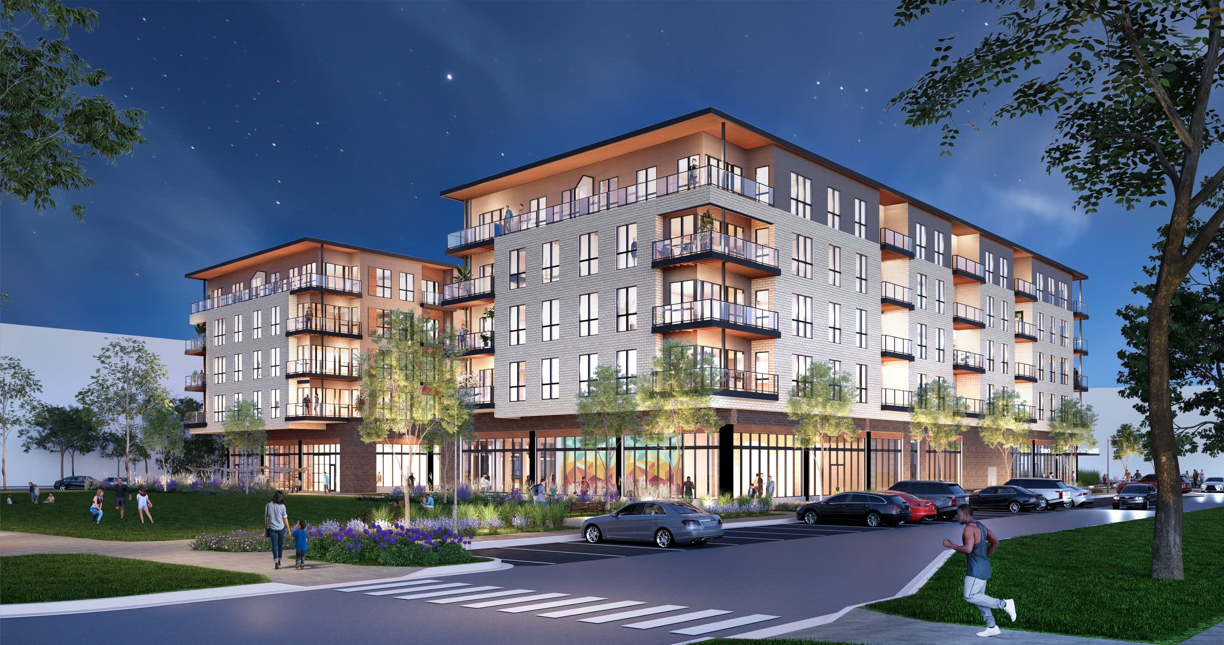 Modern multi-story apartment building at dusk with illuminated balconies, ground-floor retail, landscaping, and parked cars.