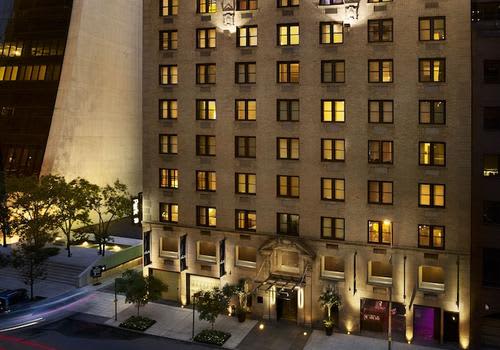 Illuminated mid-rise residential building at night with lit windows, ground-floor storefronts, and landscaped courtyard entrance.