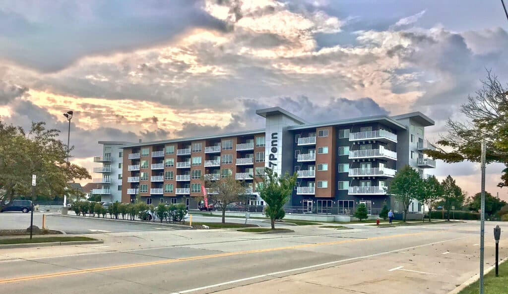 Modern multi-story apartment building with brick and blue-green facade, ground-floor retail, landscaped plaza, and street frontage.