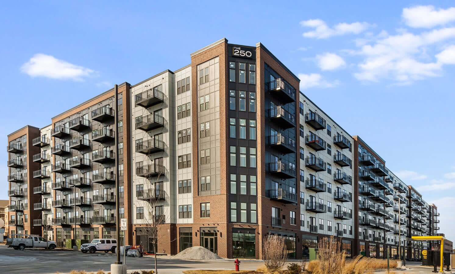 Multi-story apartment complex with brick and tan facades, metal balconies, and street-level storefronts under blue sky.