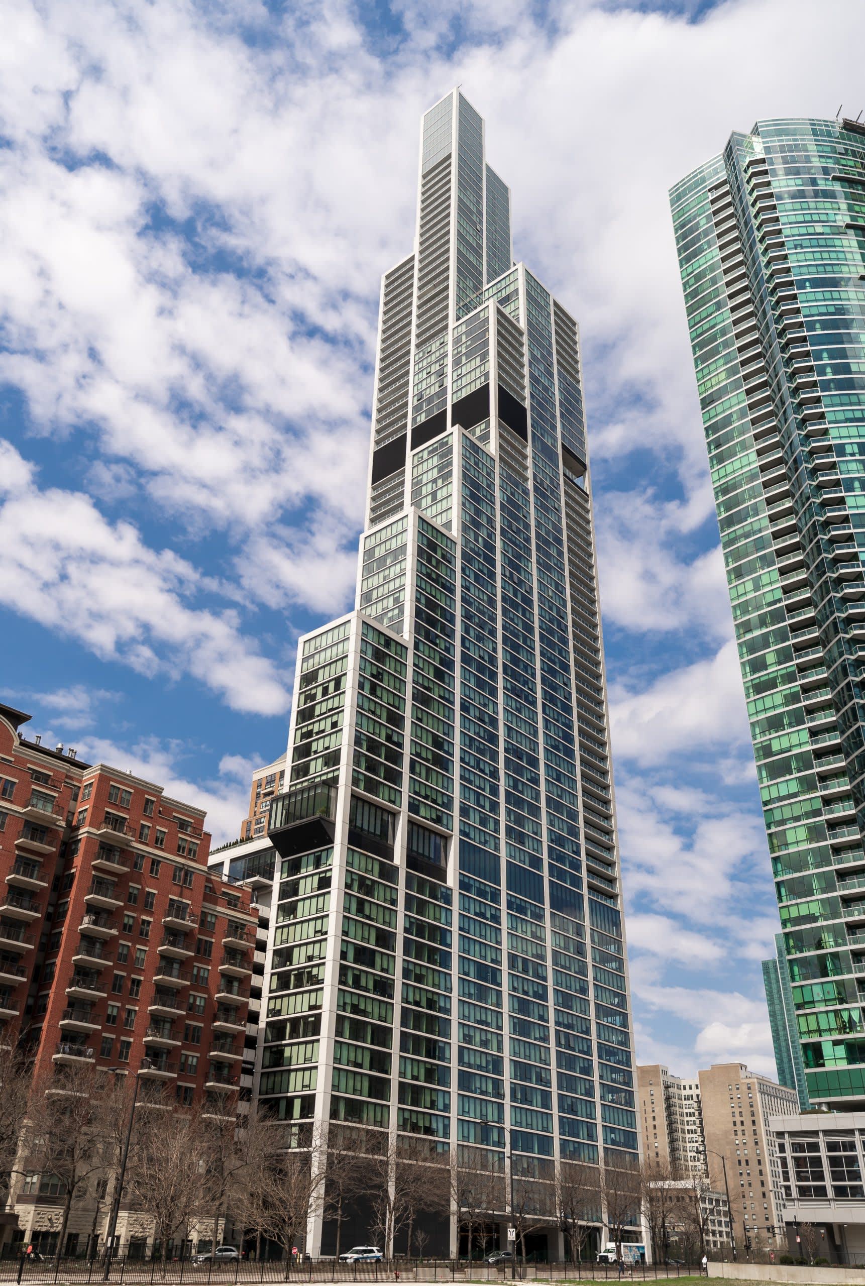 Tall residential tower with geometric white framework and green-tinted glass windows against blue sky, surrounded by other urban buildings.