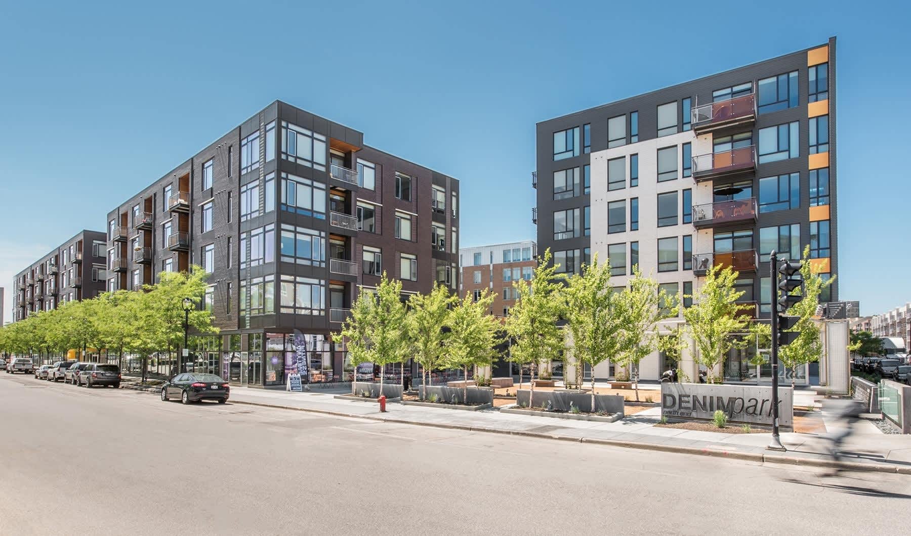 Modern apartment buildings with brick and metal facades, ground-floor retail spaces, and green trees lining the street.