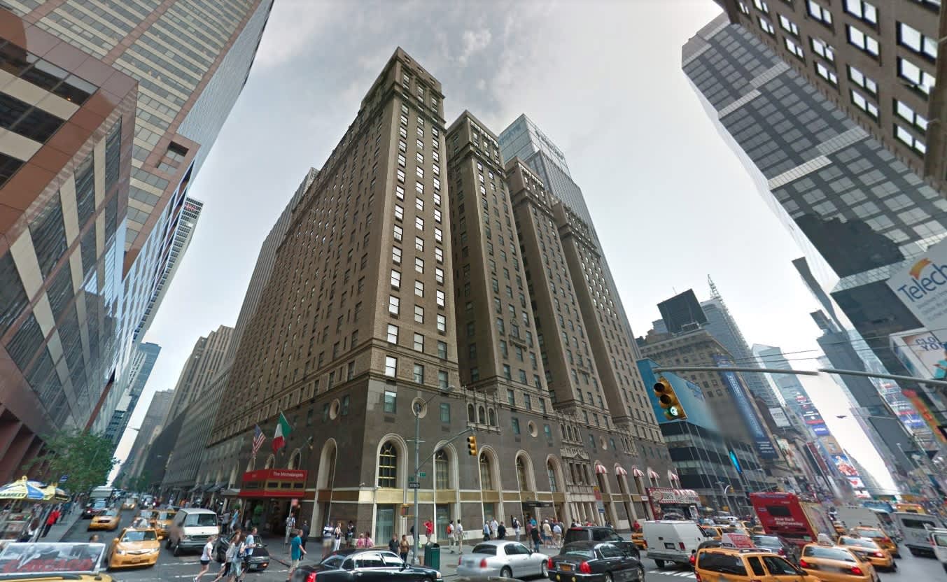 Tall historic office building with tan brick facade and arched ground-floor windows on a busy Manhattan street surrounded by skyscrapers and yellow taxis.