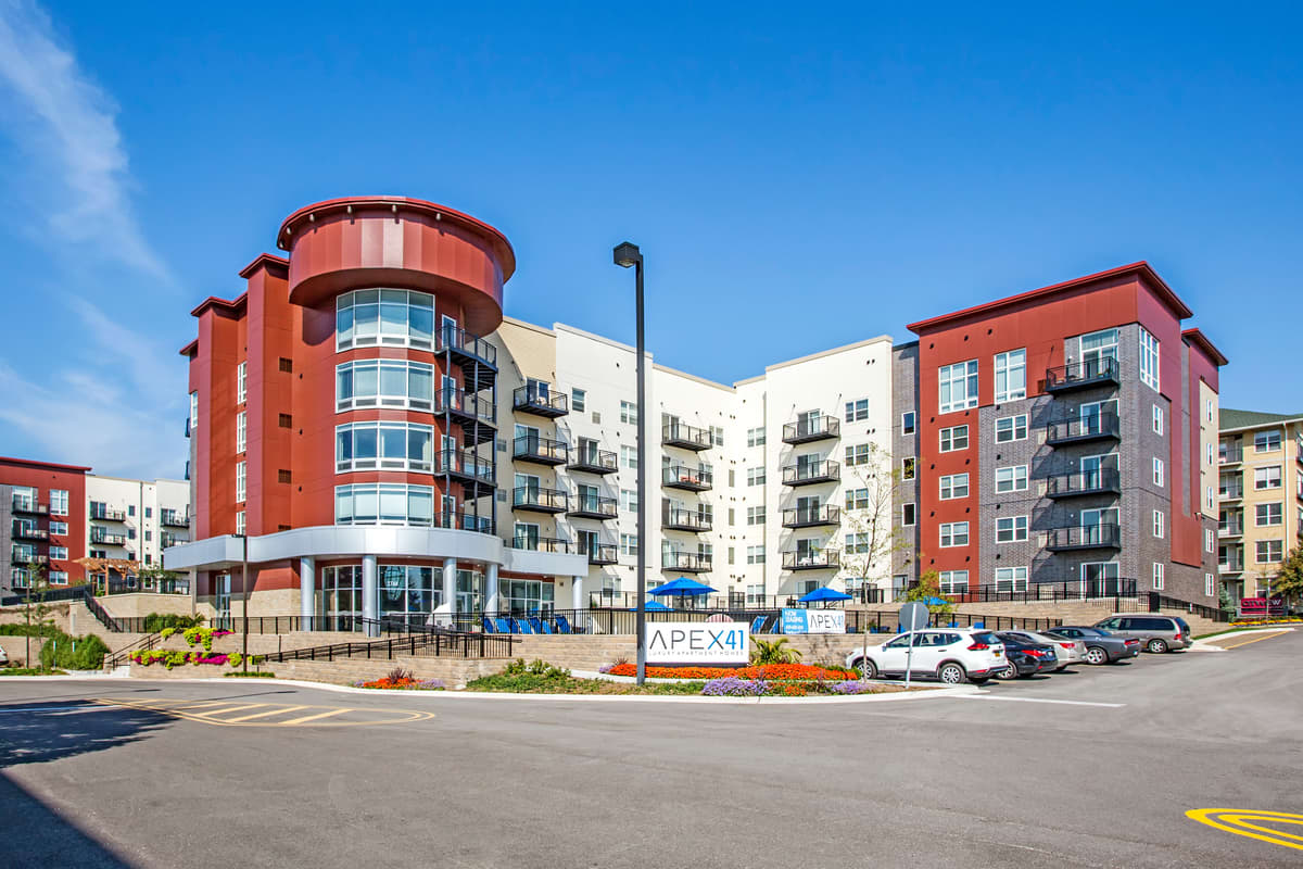 Multi-story apartment complex with red brick cylindrical tower, white and red buildings, balconies, and parking lot under clear blue sky.