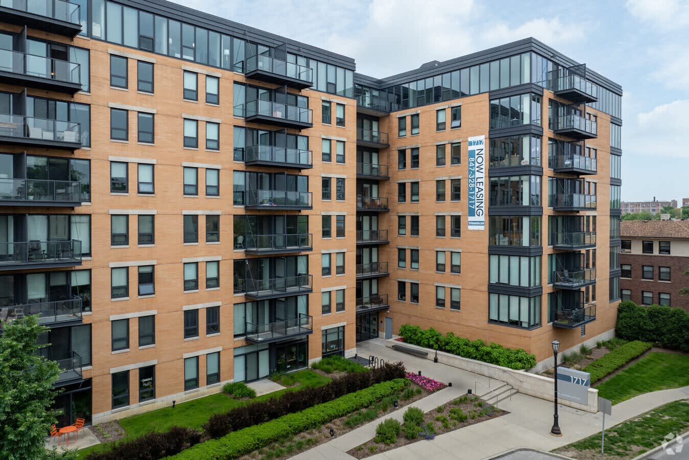 Modern multi-story apartment building with tan brick facade, dark balconies, and green courtyard landscaping.