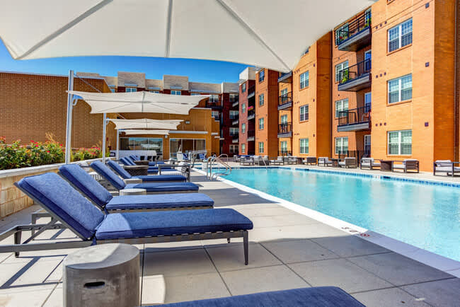 Resort-style swimming pool with blue lounge chairs, white shade structures, and multi-story brick apartment buildings in background.