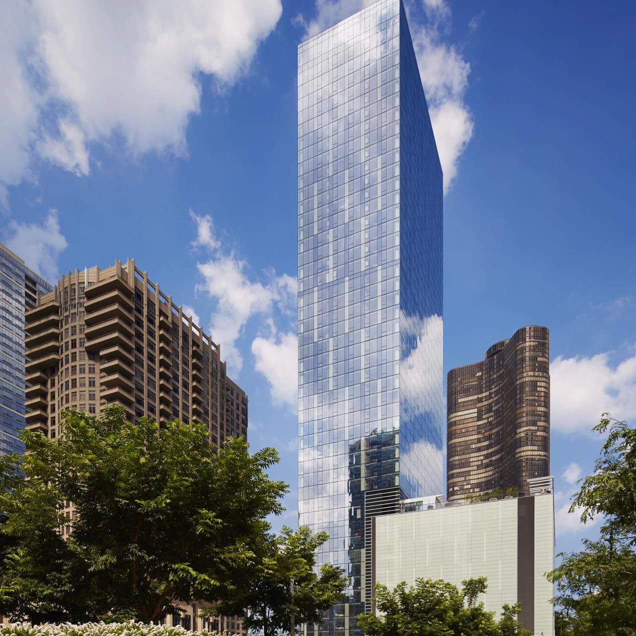 Tall glass and steel high-rise tower with reflective windows, flanked by older brick residential buildings and green trees under blue sky.