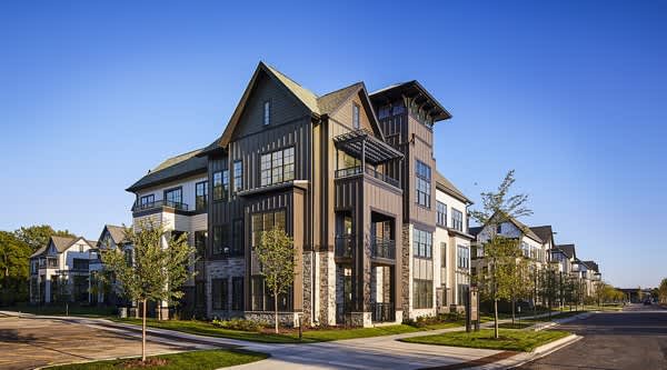 Multi-story residential building with black and tan exterior, large windows, and surrounding young trees on a clear day.
