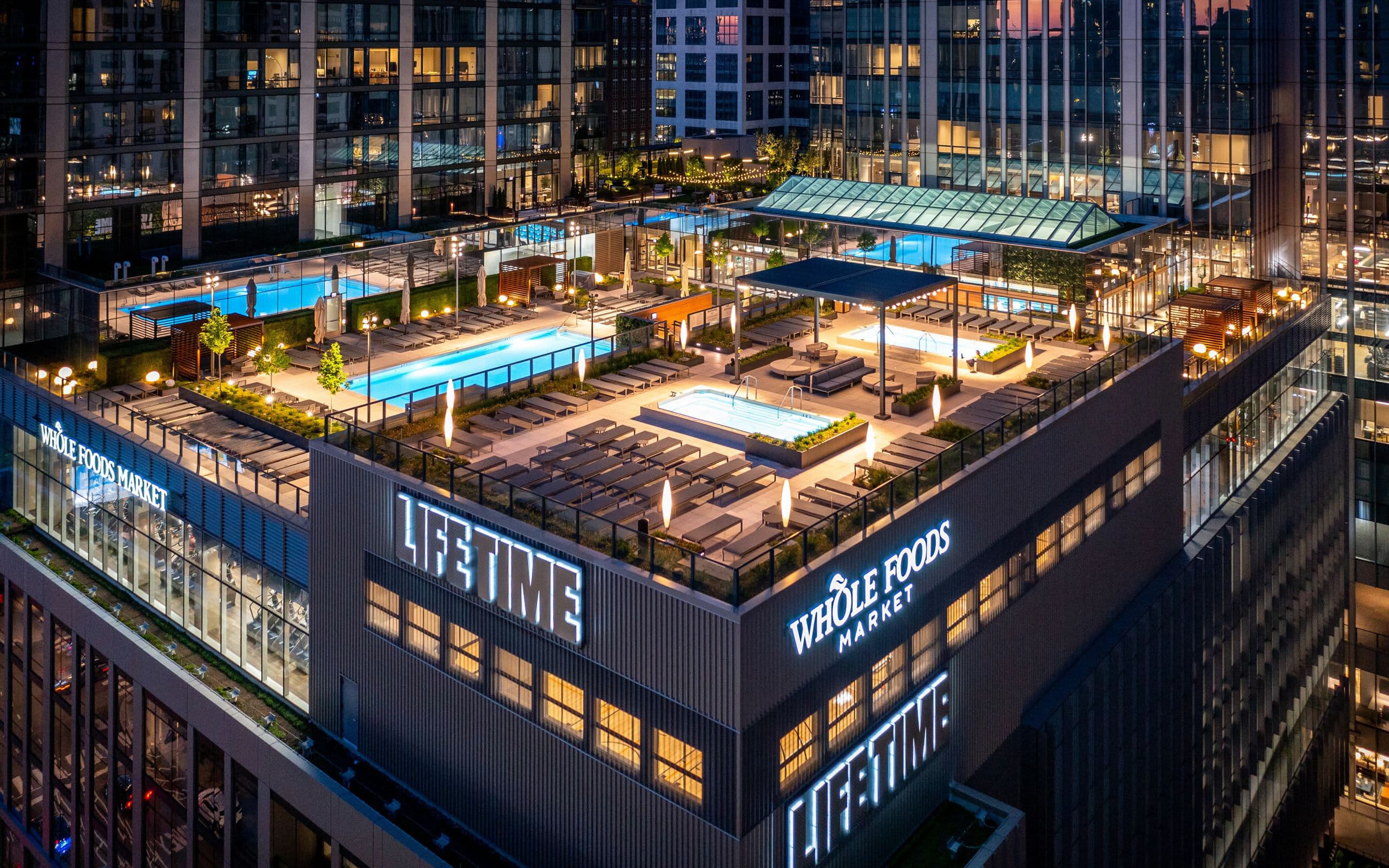 Aerial view of illuminated rooftop deck with two blue pools, lounge seating, shade structures, and Whole Foods Market signage surrounded by Chicago high-rises at dusk.