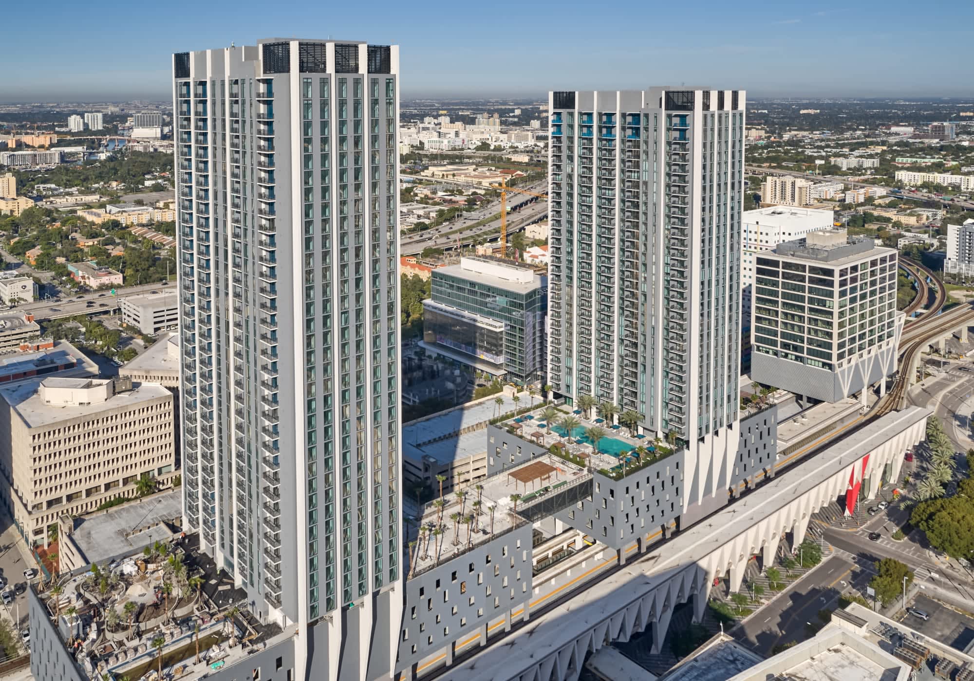 Two tall residential towers with white and glass facades overlooking a central courtyard with pool and landscaping, surrounded by urban infrastructure and highways.