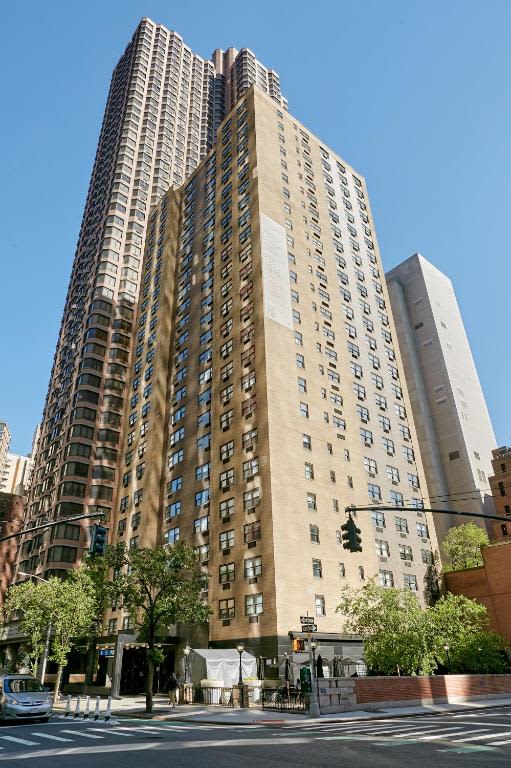 Tall residential building with tan and dark brick facade, regular window grid, street-level entrance, and surrounding city buildings under clear sky.