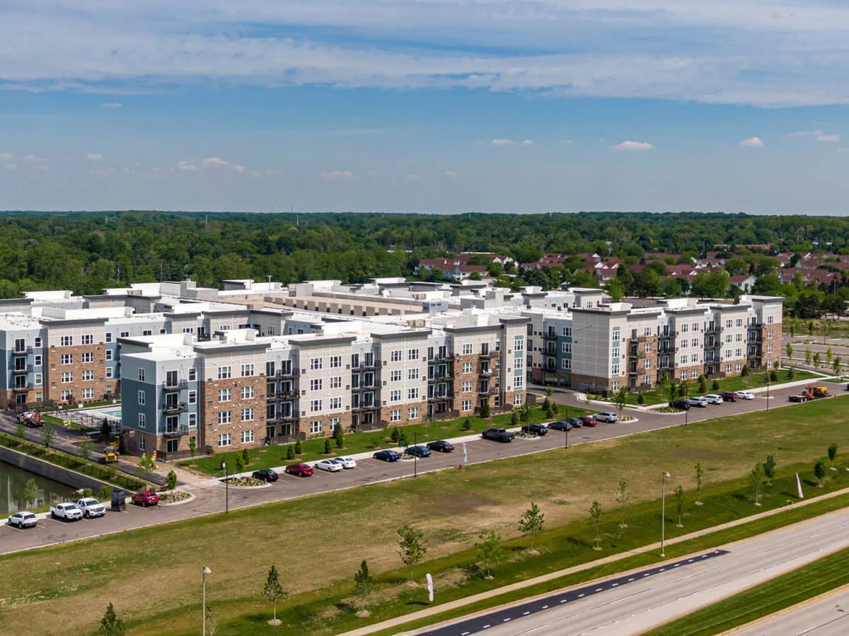 Aerial view of multi-story apartment complex with brick and tan facades, parking lot, green landscaping, and wooded residential area beyond.