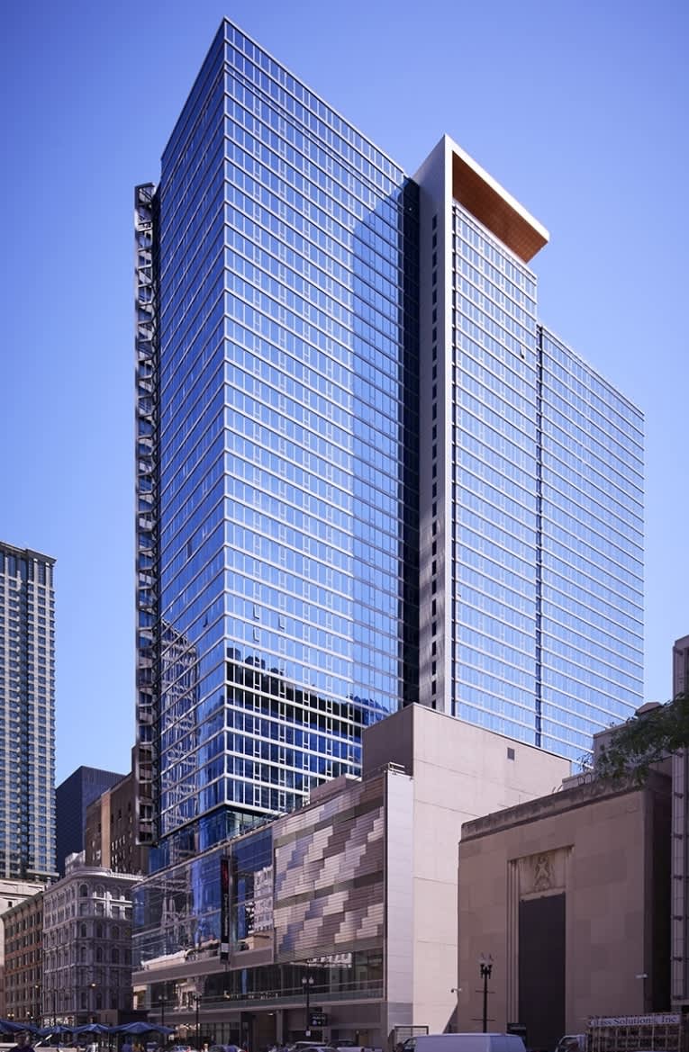 Modern high-rise residential tower with blue-tinted glass facade and copper architectural accent, situated above a cream-colored base building in an urban downtown setting.