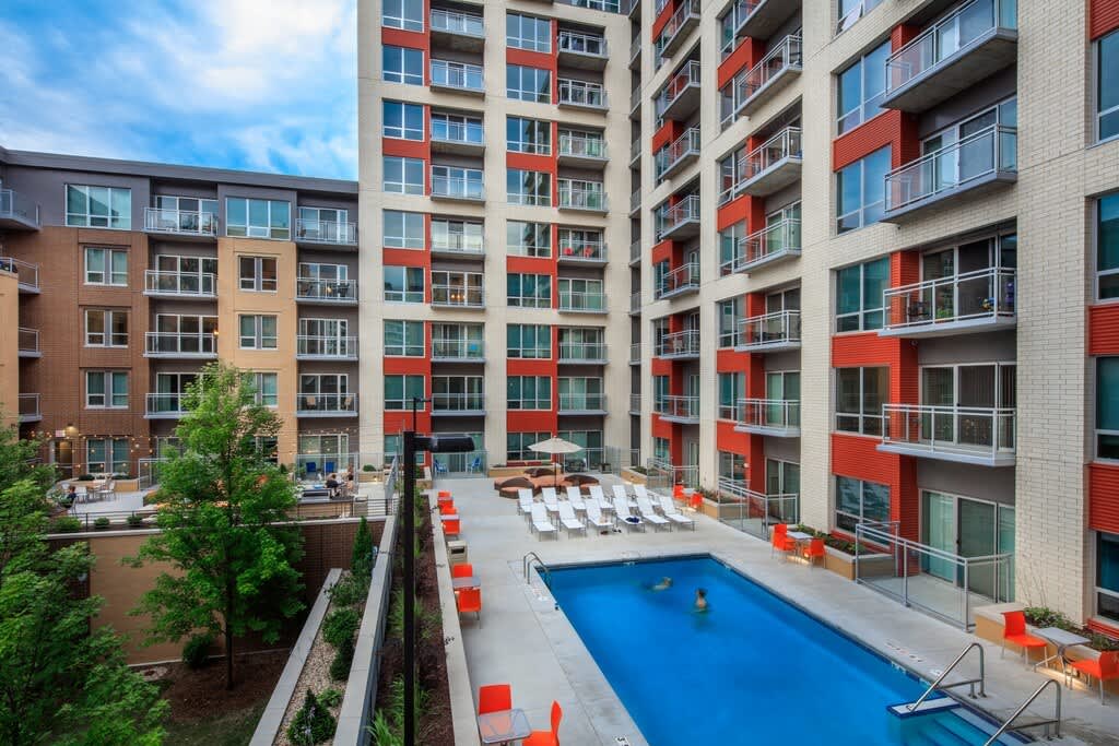 Courtyard pool area with blue water, white lounge chairs, red accent seating, and multi-story residential buildings with red and tan facades.