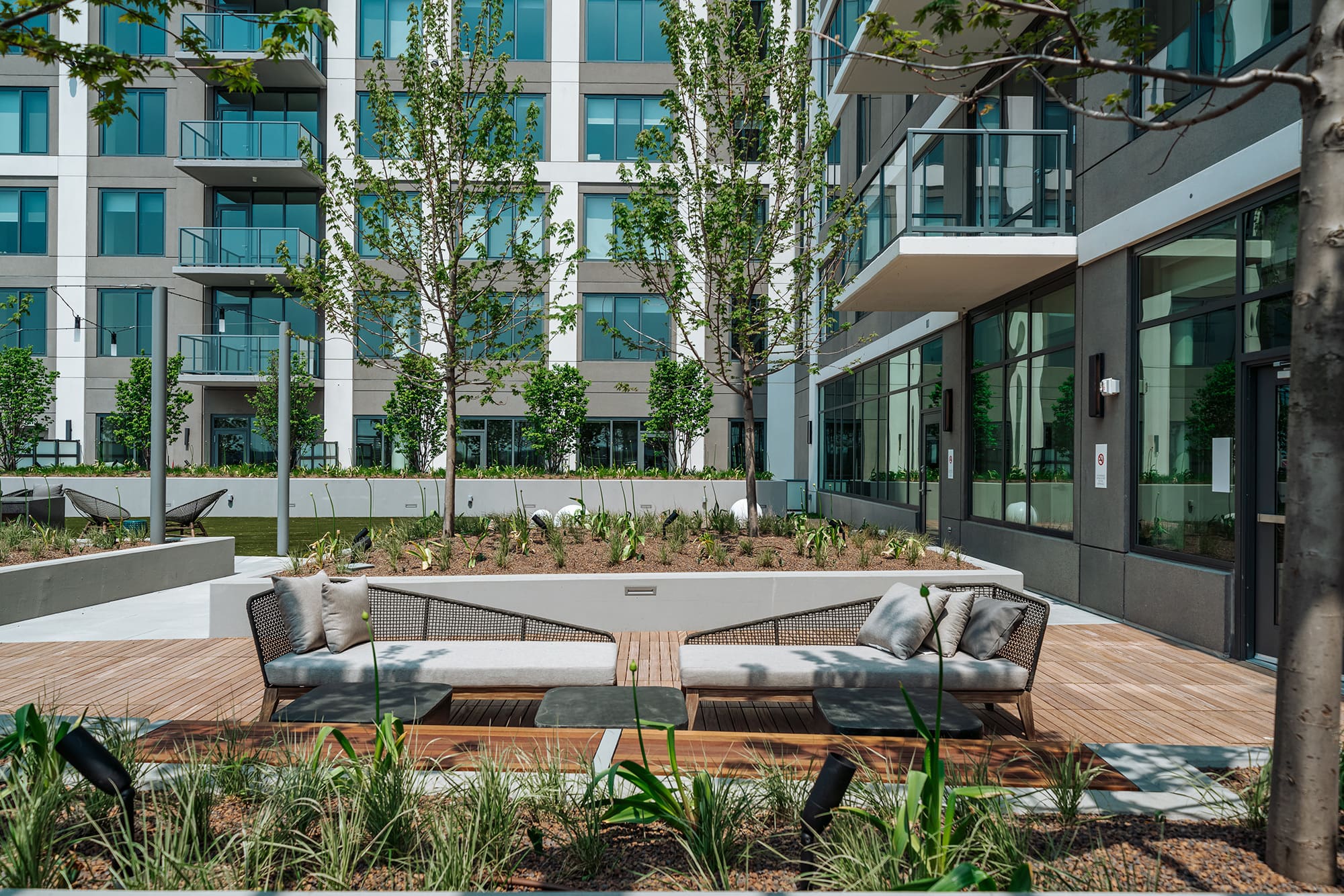 Outdoor courtyard with two woven lounge chairs, wooden deck, young trees, and modern multi-story residential buildings with glass balconies.
