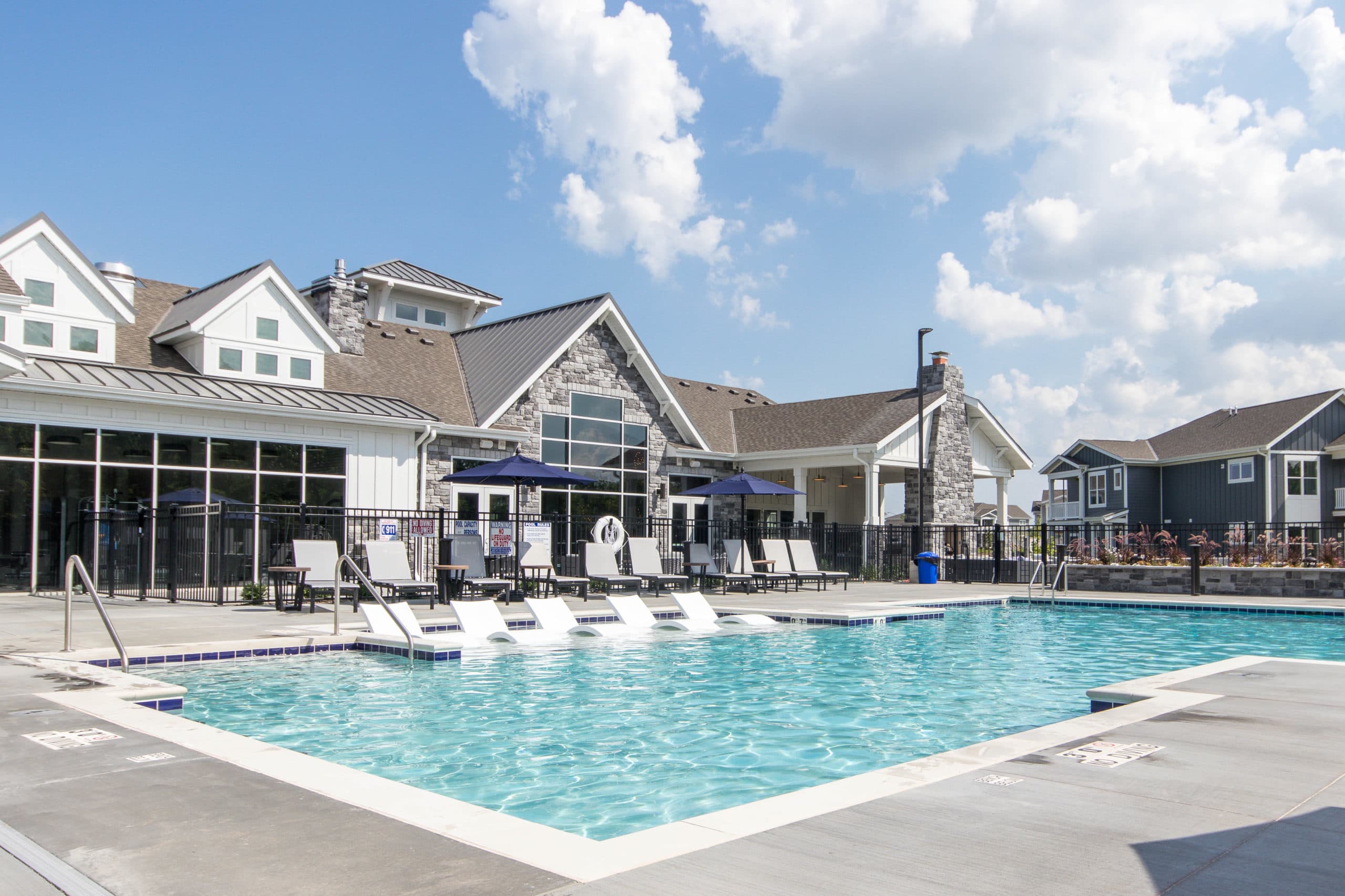 Resort-style swimming pool with white lounge chairs, concrete deck, and modern clubhouse building with stone facade and metal roof.