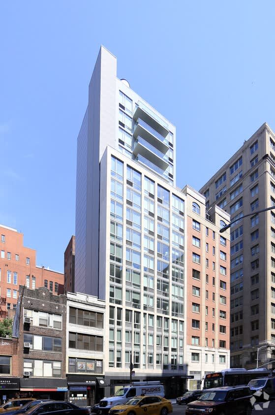 Modern white and glass residential building with multiple floors and balconies, surrounded by older brick buildings and street-level storefronts.