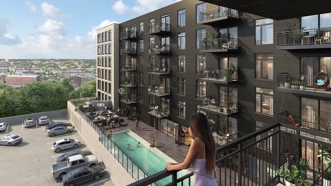 Woman on balcony overlooking rooftop pool, modern dark apartment building, and parking lot with city views.