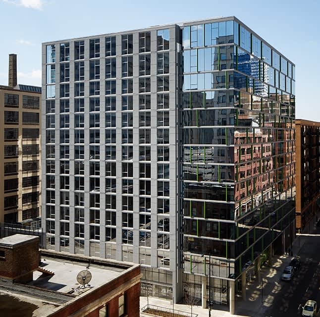 Modern multi-story residential building with extensive glass windows and metal framing, adjacent to historic brick buildings in an urban setting.