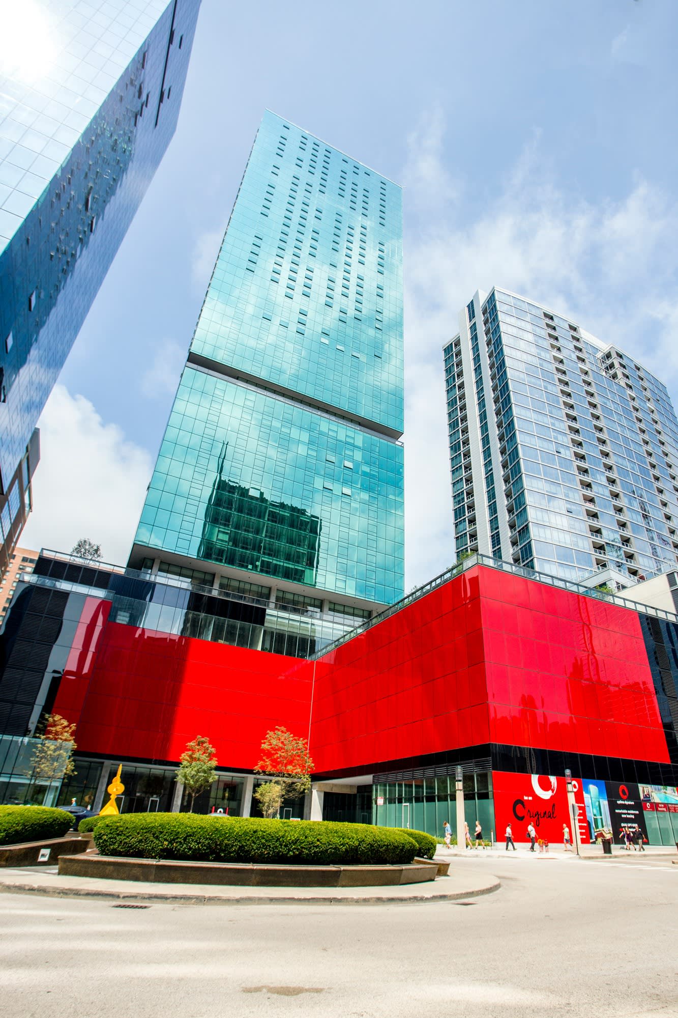 Modern high-rise residential towers with glass and turquoise facades, red cultural building below, landscaped plaza with greenery.