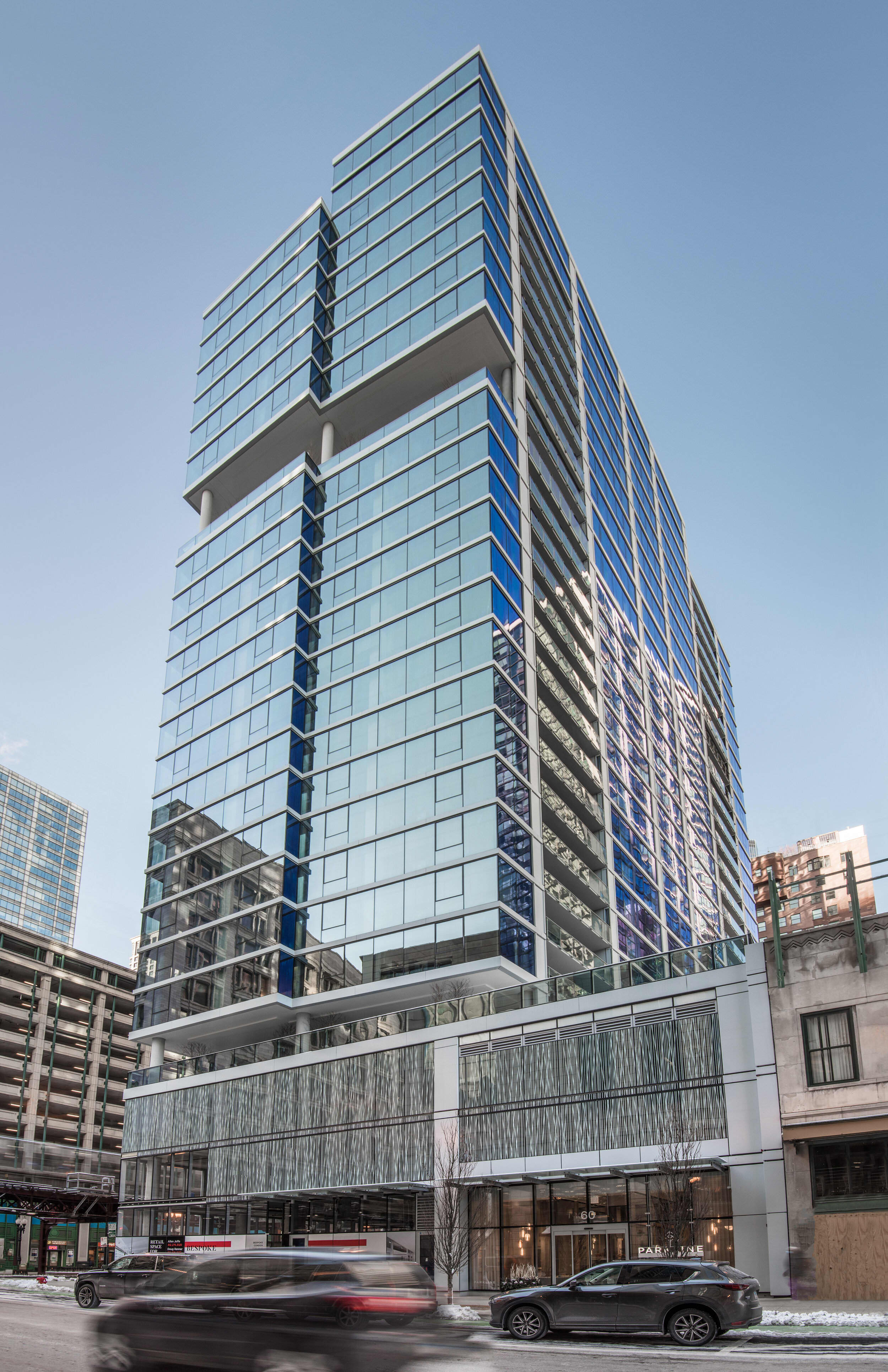 Modern high-rise residential building with glass facade, horizontal balconies, and ground-level retail space in urban setting.