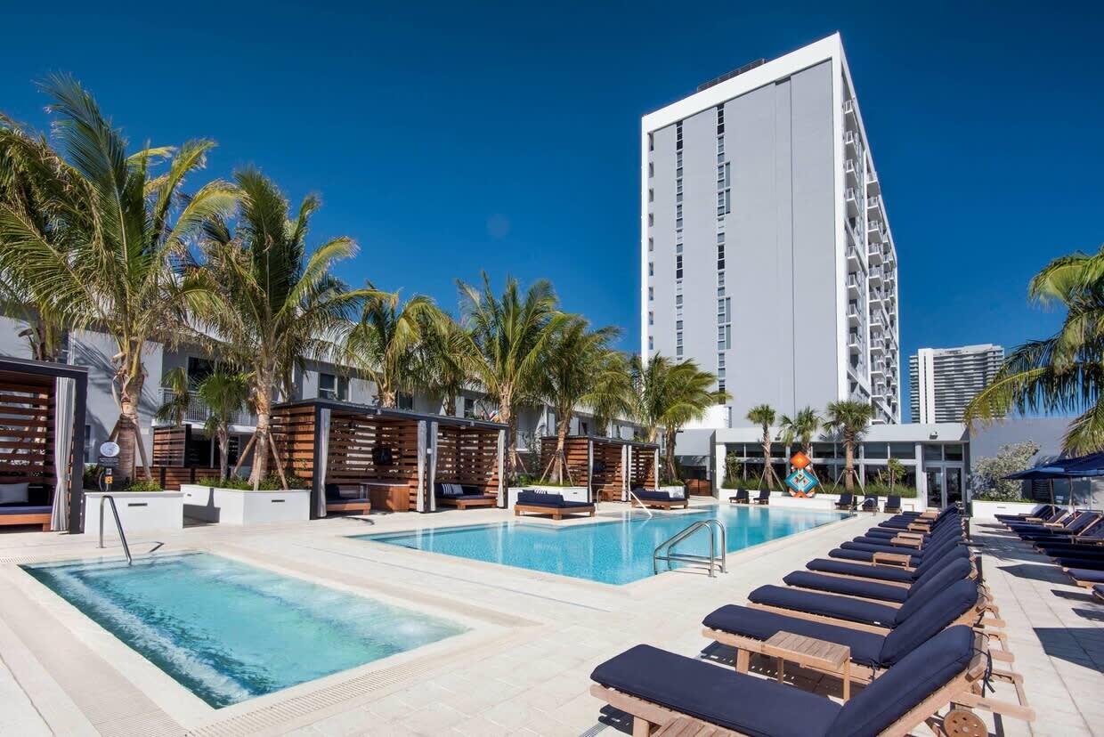 Resort pool deck with two pools, wooden lounge chairs, palm trees, and white residential tower under clear blue sky.