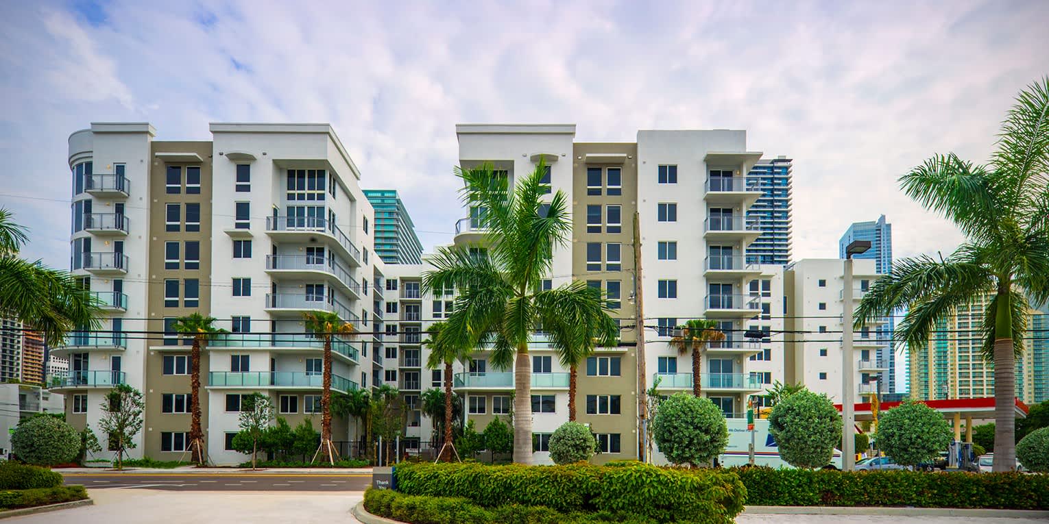 Multi-story residential buildings with cream and glass facades, palm trees, and manicured green landscaping in foreground.