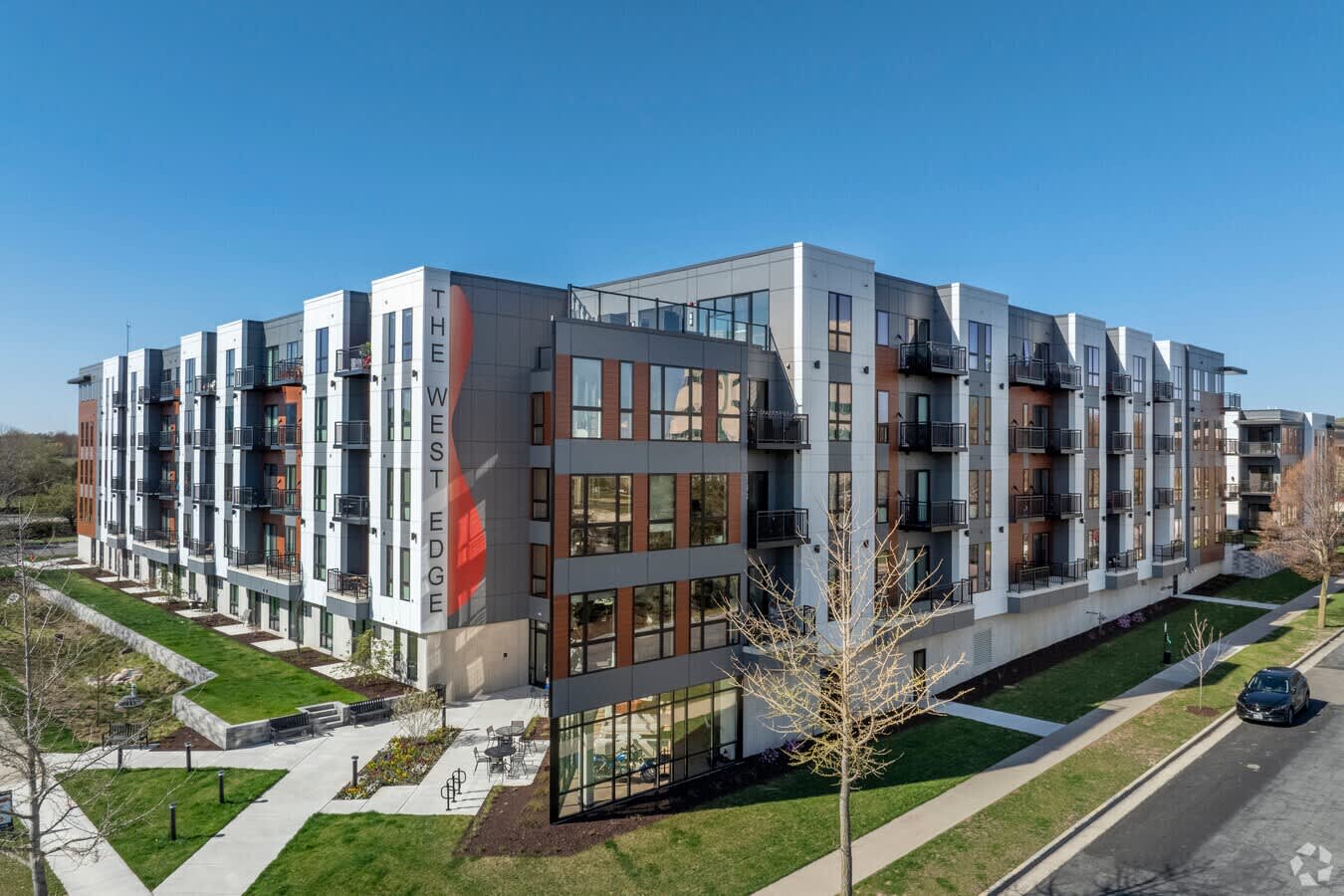 Modern four-story apartment building with white, gray, and brick exterior, multiple balconies, large windows, and manicured green lawn.