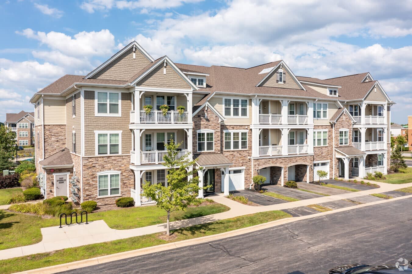 Multi-story residential building with brick and tan siding, white trim, covered balconies, and attached garages along a tree-lined street.