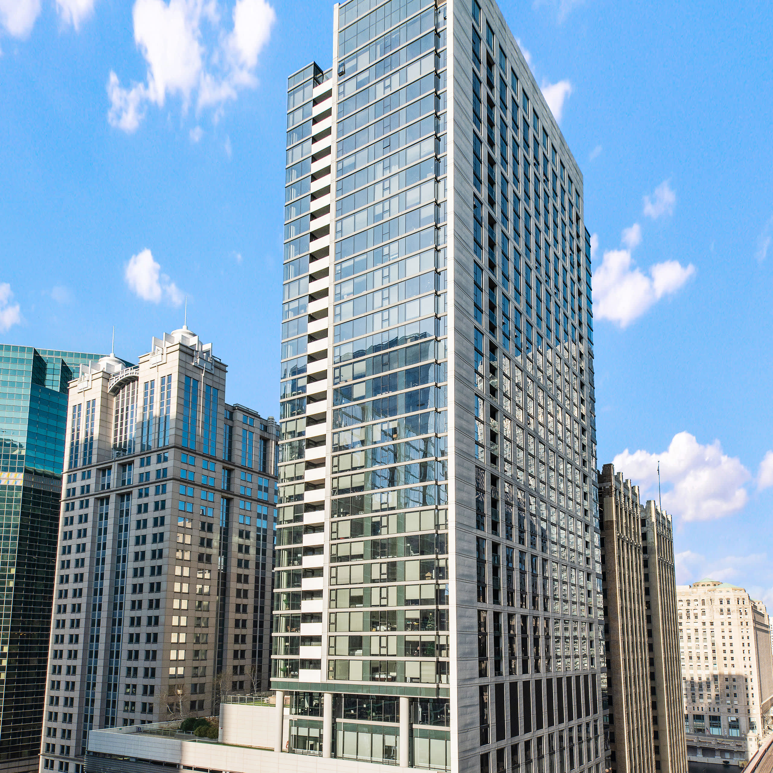 Modern high-rise residential tower with glass and metal facade among surrounding downtown Chicago buildings under blue sky.