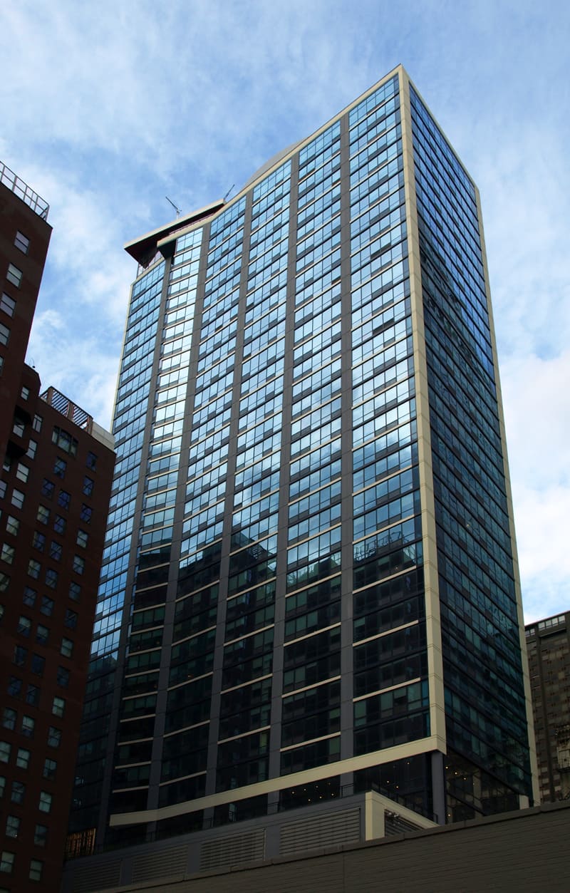 Tall residential high-rise building with gridded glass windows and tan metal framing against blue sky.