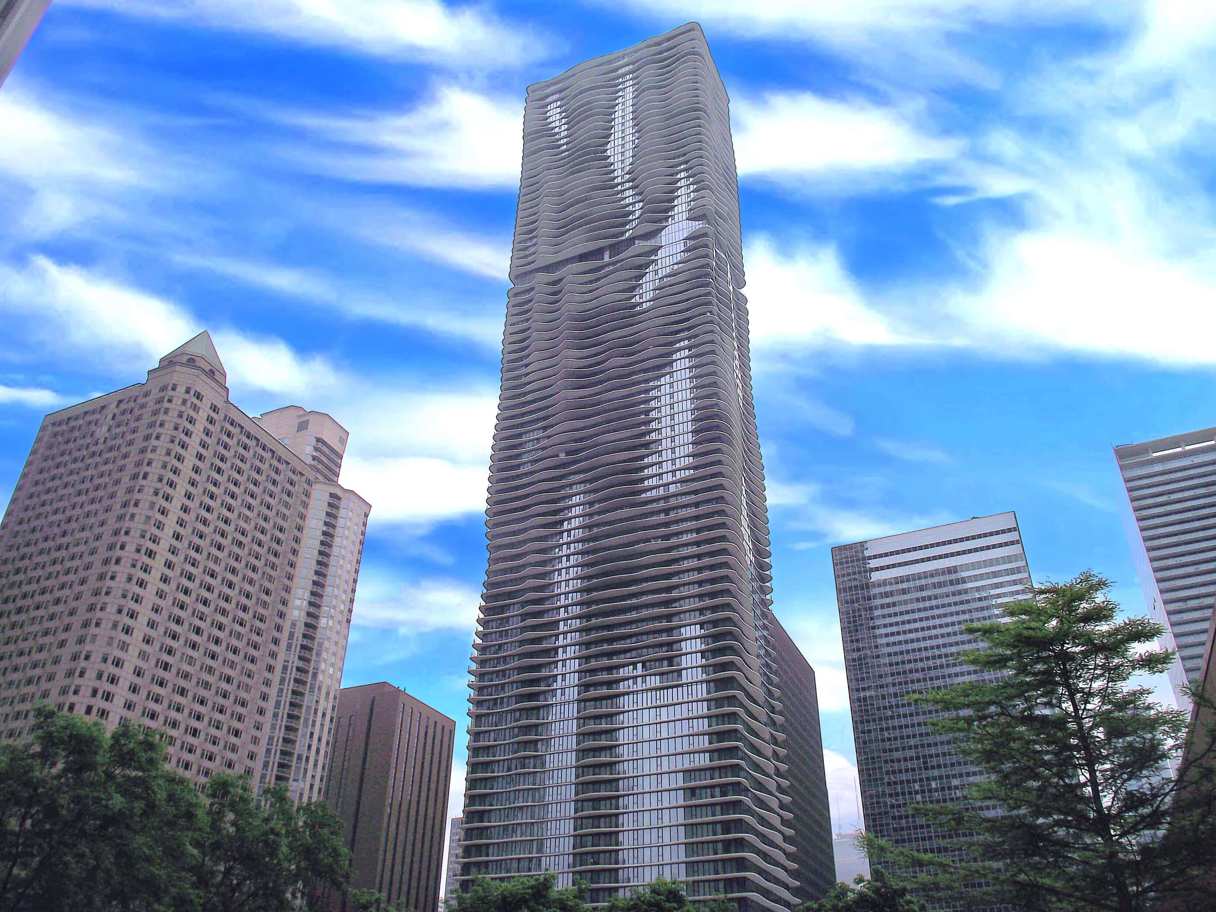 Tall residential tower with curved balconies among surrounding downtown buildings under blue sky.