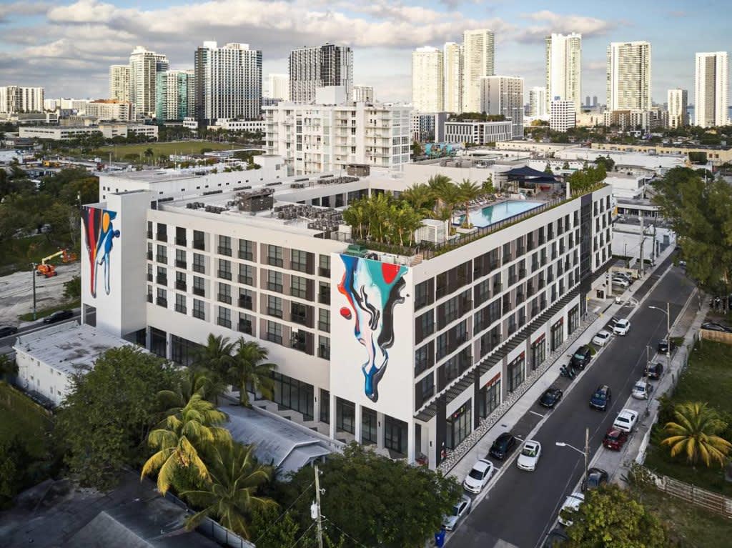 Modern white residential building with colorful mural art, rooftop pool with palm trees, and Miami downtown skyline in background.