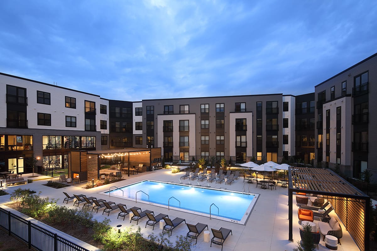 Courtyard with lit swimming pool, lounge chairs, umbrellas, and multi-story residential buildings at dusk.
