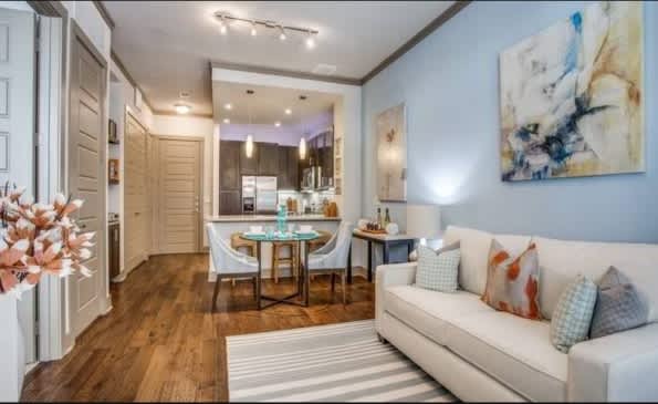 Open-concept apartment with cream sofa, round dining table, dark kitchen with island, hardwood floors, and blue accent wall.