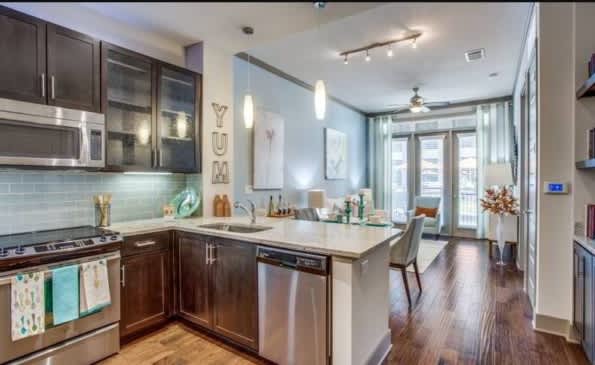 Modern kitchen with dark wood cabinetry, stainless steel appliances, white quartz island, glass backsplash, and open living space beyond.